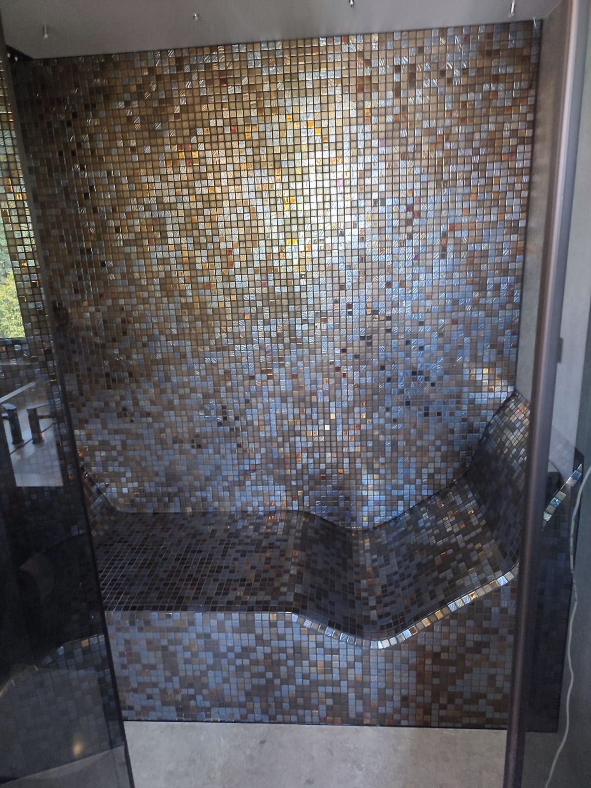 Mozaika w odcieniach złota i błękitu zdobi ścianę prysznica z wbudowanym siedziskiem o zaokrąglonych krawędziach, widoczne przez szklaną taflę.