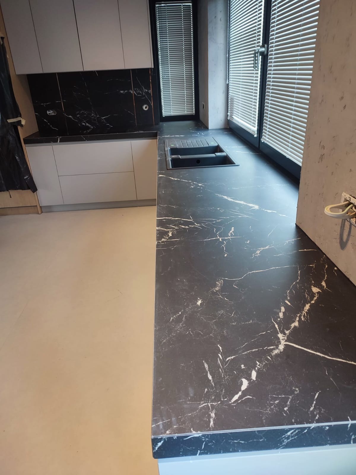 Nowoczesna kuchnia w trakcie wykańczania: czarny blat z marmurowym wzorem, wbudowany zlewozmywak, białe szafki i czarne płytki na ścianie, okno z żaluzjami, surowe ściany.