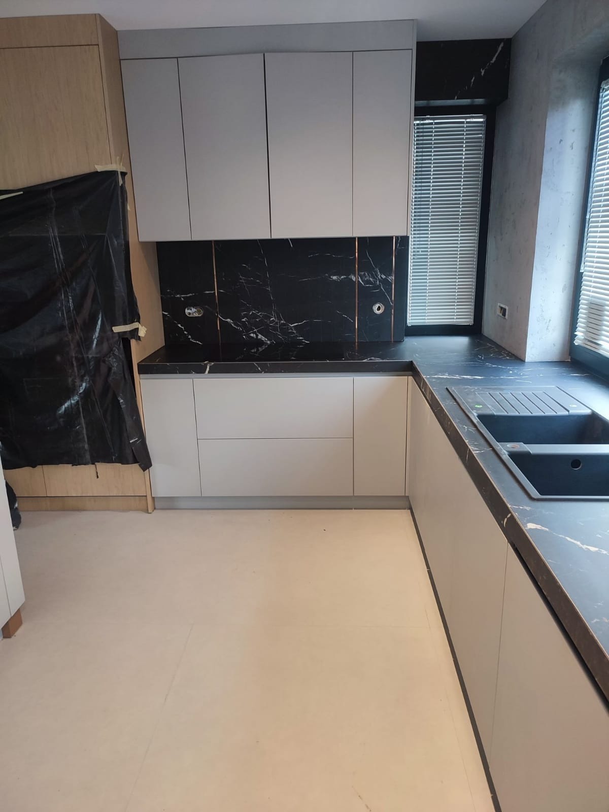 Nowoczesna kuchnia w trakcie wykańczania: szare szafki, czarny blat i backsplash z marmurowym wzorem, okno z żaluzjami, podłoga zabezpieczona folią, widoczne gniazdka elektryczne.