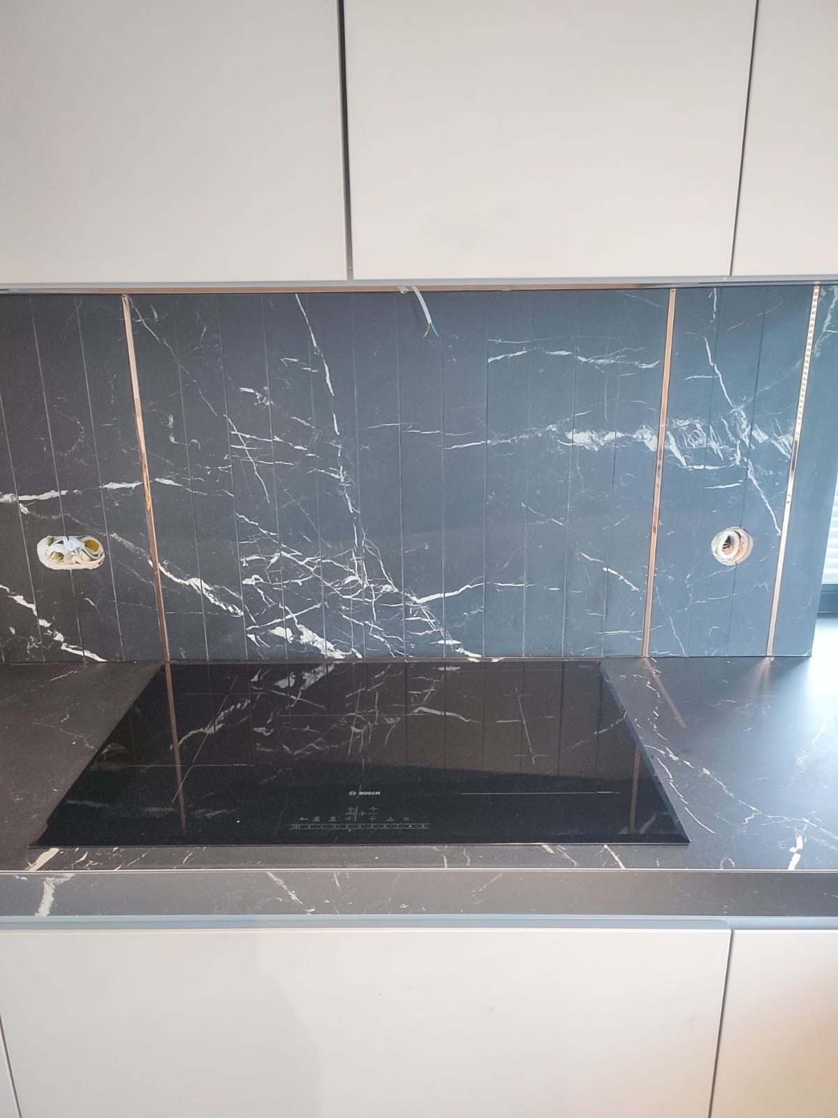 Czarne płytki imitujące marmur z miedzianymi listwami na ścianie nad czarną płytą indukcyjną Bosch, widoczne gniazdka elektryczne w trakcie montażu.