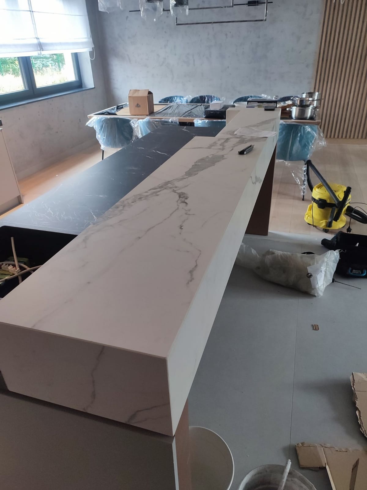 Montaż blatu kuchennego z białego marmuru z szarymi żyłami, widoczny fragment czarnego blatu obok, w tle zakryte meble i żółty odkurzacz.