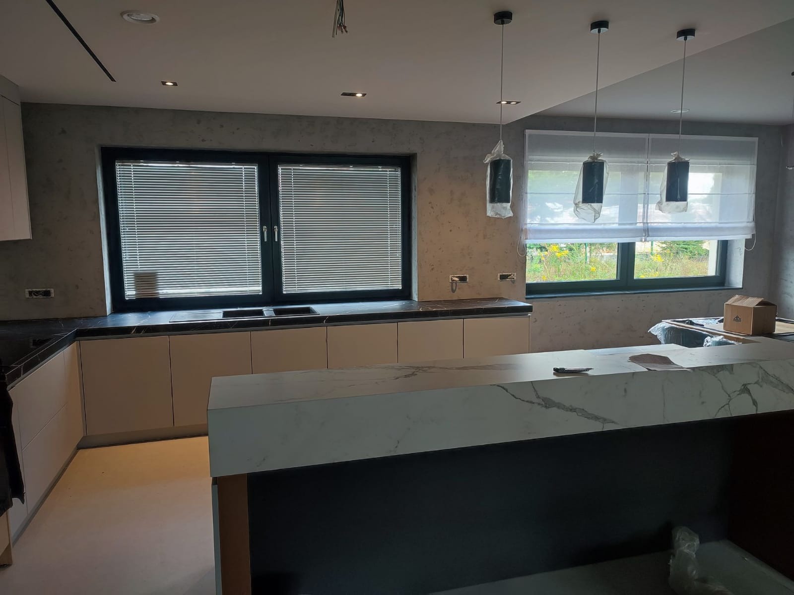 Wnętrze kuchni w trakcie wykańczania, widoczne beżowe szafki z czarnym blatem, marmurowy blat wyspy, okna z żaluzjami i roletami rzymskimi oraz lampy wiszące w folii ochronnej.