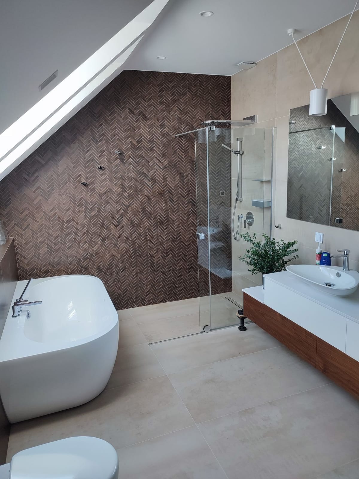 Nowoczesna łazienka na poddaszu z brązową ścianą w jodełkę, białą wanną wolnostojącą, kabiną prysznicową typu walk-in i umywalką nablatową z szafką z drewnianymi akcentami.