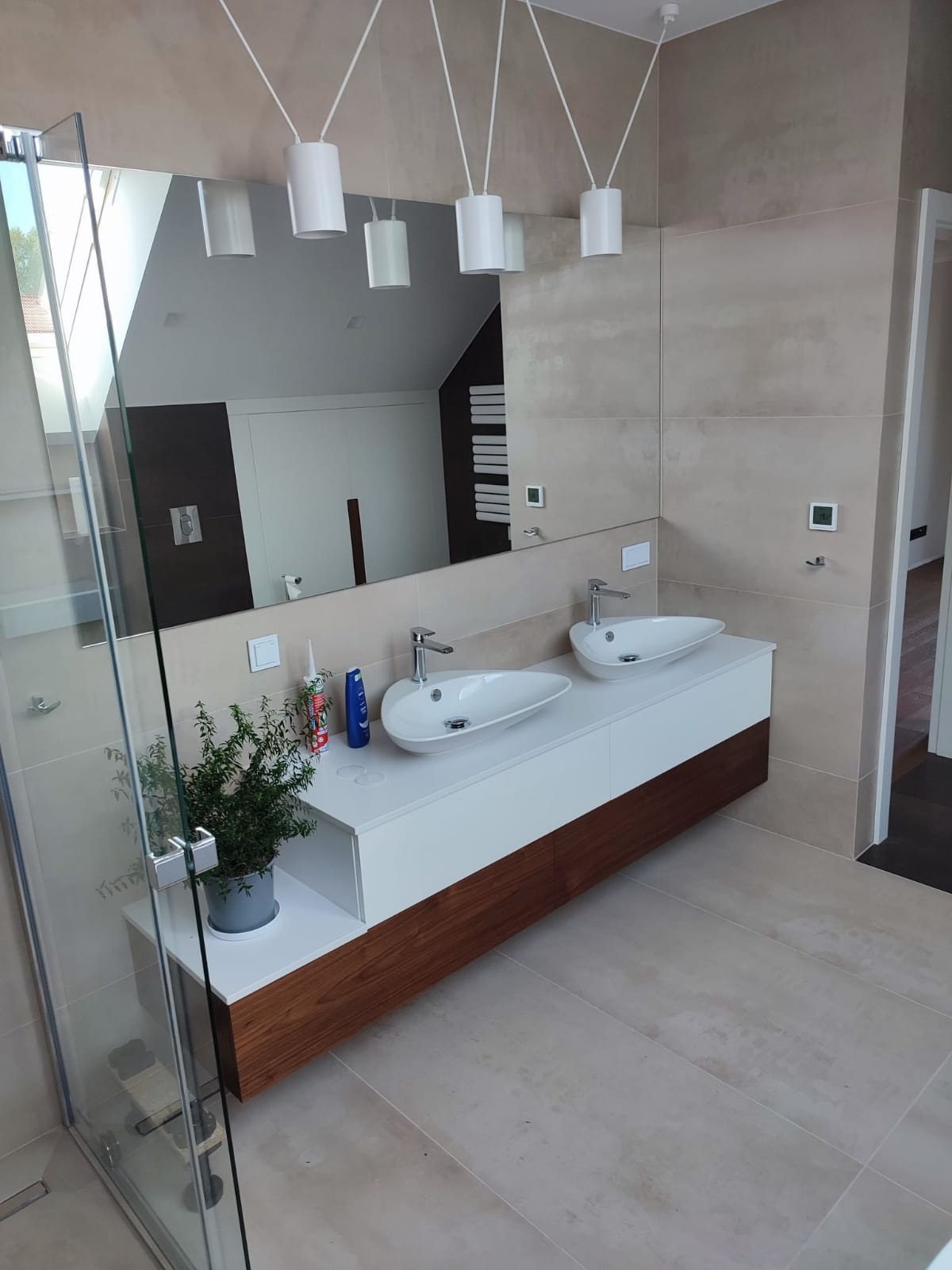 Nowoczesna łazienka z dwoma owalnymi umywalkami nablatowymi, szafką z drewnianym frontem, dużym lustrem i wiszącymi lampami cylindrycznymi; na blacie widoczne akcesoria montażowe i roślina w doniczce.