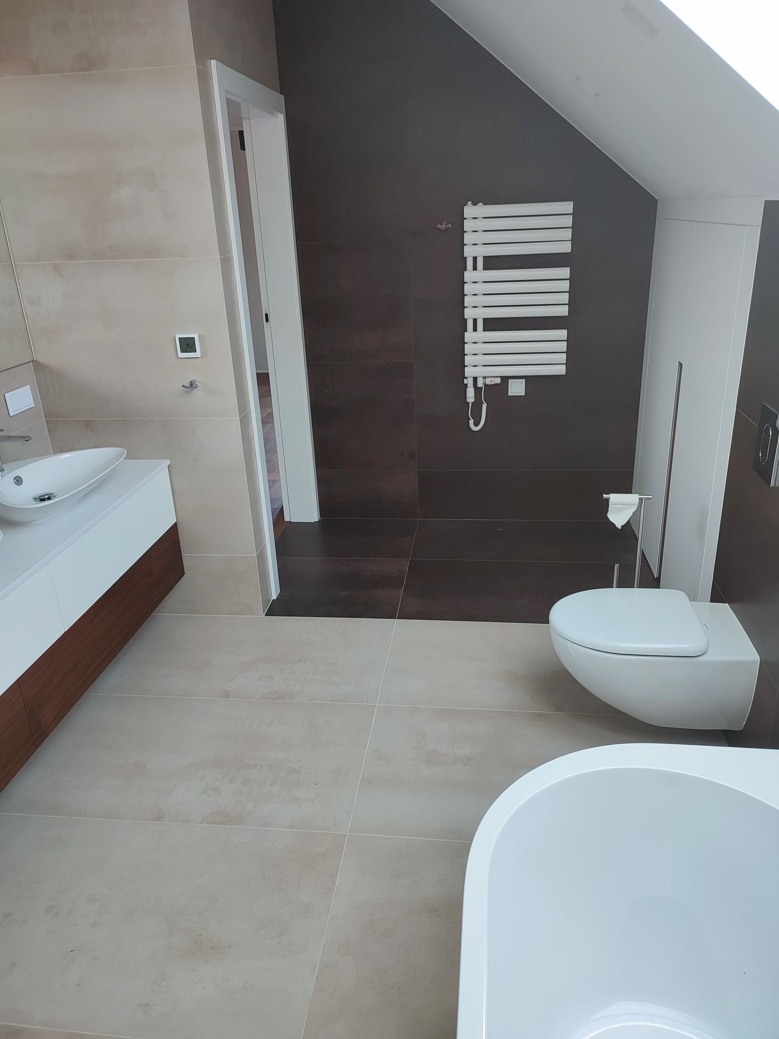 Nowoczesna łazienka na poddaszu z beżowymi płytkami, brązową ścianą, białą armaturą, grzejnikiem drabinkowym i umywalką nablatową z szafką z drewnianym akcentem.