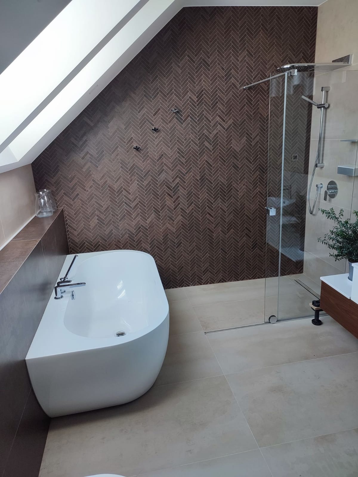 Nowoczesna łazienka na poddaszu z wolnostojącą wanną, kabiną prysznicową ze szklanymi drzwiami i ścianą wyłożoną brązowymi płytkami ułożonymi w jodełkę. Uwagę przyciągają czarne haczyki na ręczniki...