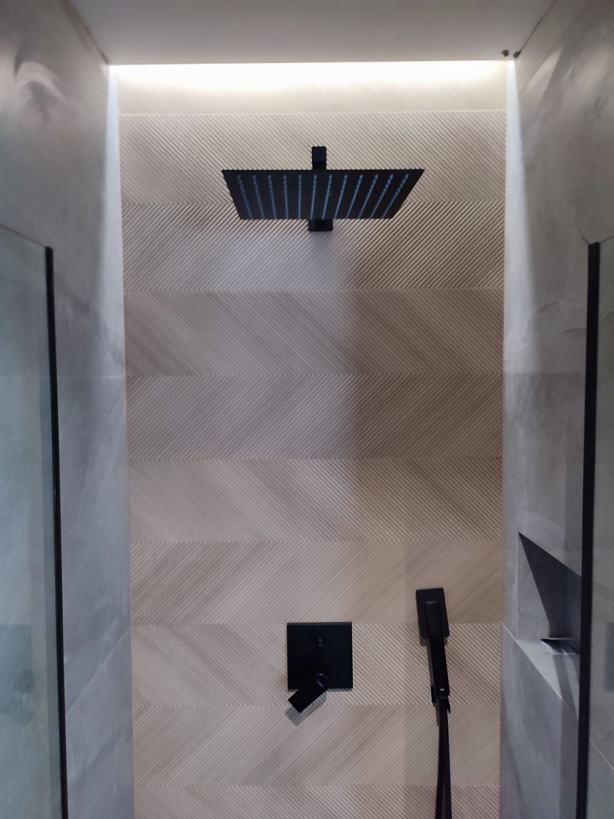 Nowoczesna kabina prysznicowa z deszczownicą w kolorze czarnym, geometryczna tekstura płytek na ścianie, oświetlenie LED pod sufitem, minimalistyczny design.