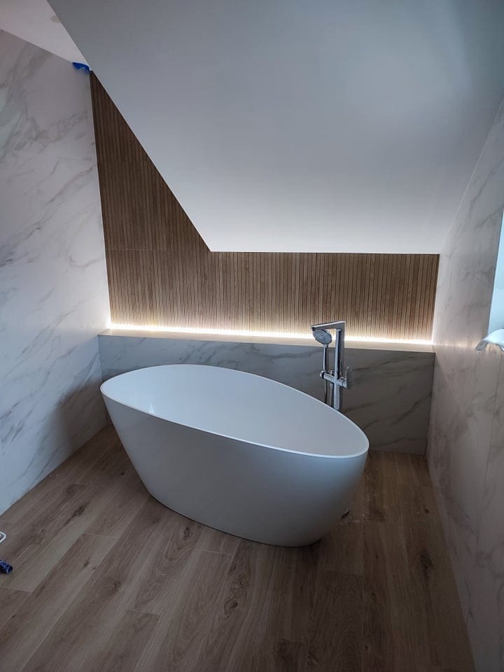 Nowoczesna łazienka na poddaszu z wolnostojącą białą wanną, marmurowymi płytkami na ścianach, drewnianą boazerią ze zintegrowanym oświetleniem LED i podłogą imitującą drewno.