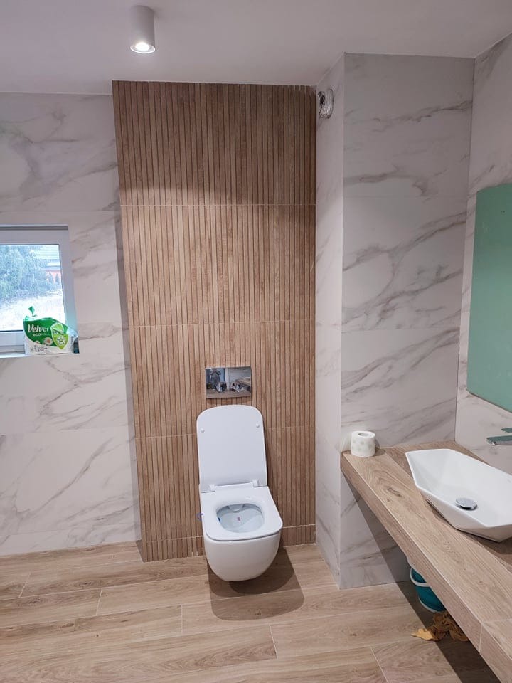 Nowoczesna łazienka w Katowicach z marmurowymi płytkami, drewnopodobną podłogą i ścianą z pionowych paneli, wisząca toaleta z podtynkowym systemem spłukiwania, umywalka nablatowa na drewnianym blacie.
