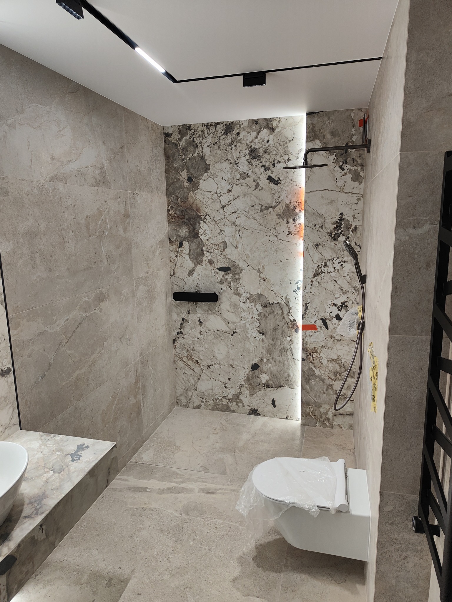 Nowoczesna łazienka w trakcie wykańczania, z marmurowymi płytkami na ścianach, podwieszaną toaletą w folii ochronnej i czarną armaturą, oświetlona listwą LED.