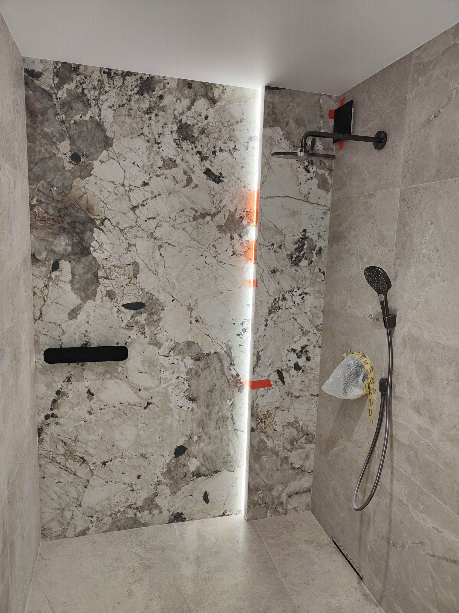 Nowoczesna kabina prysznicowa z marmurowymi płytami, czarną armaturą i podświetleniem LED w szczelinie między płytami, widoczna folia ochronna na słuchawce prysznicowej.