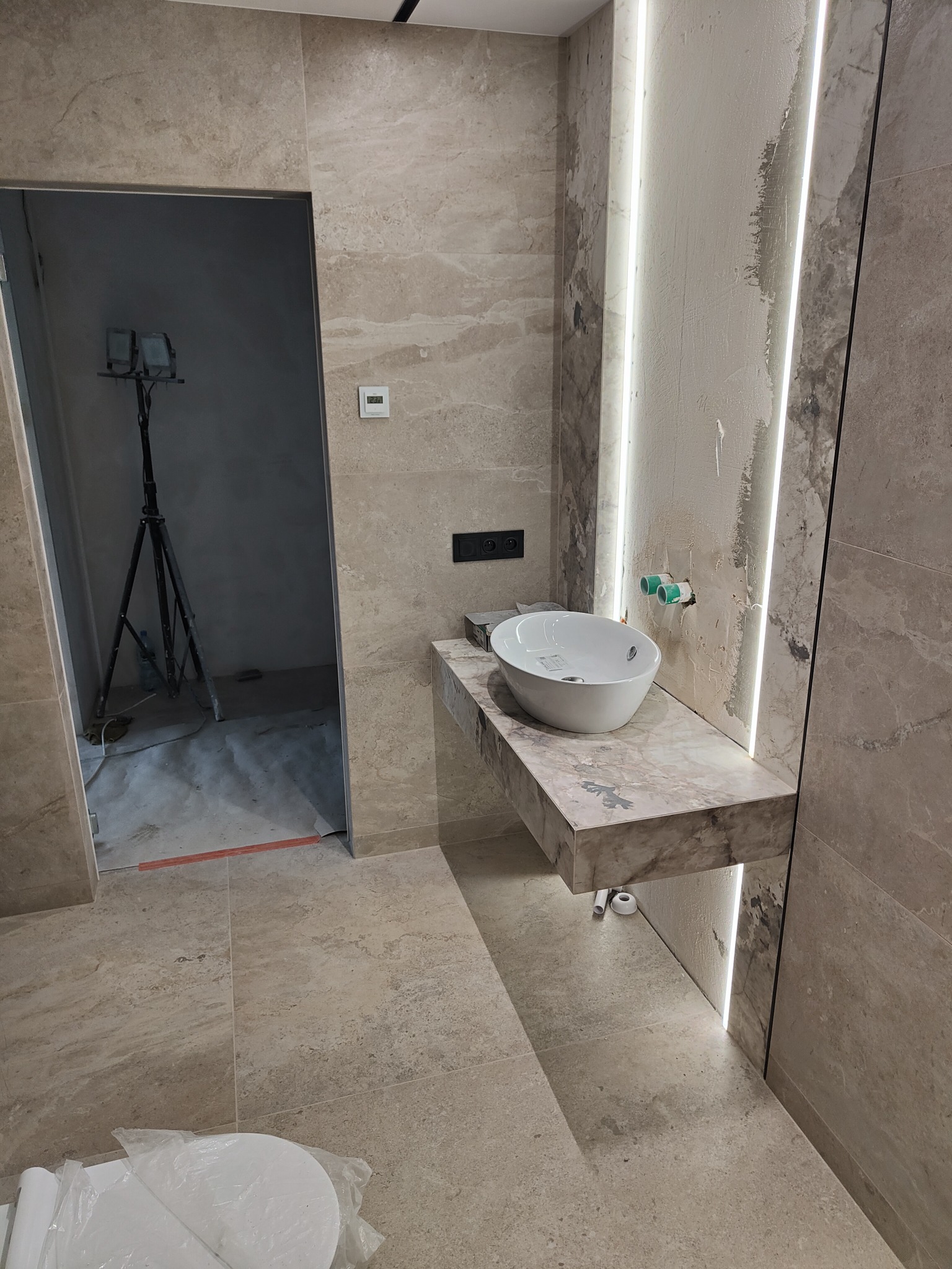 Łazienka w trakcie wykańczania; widoczna umywalka nablatowa na marmurowym blacie z podświetleniem LED, surowe ściany z instalacją wodną, płytki podłogowe i oświetlenie budowlane w tle.