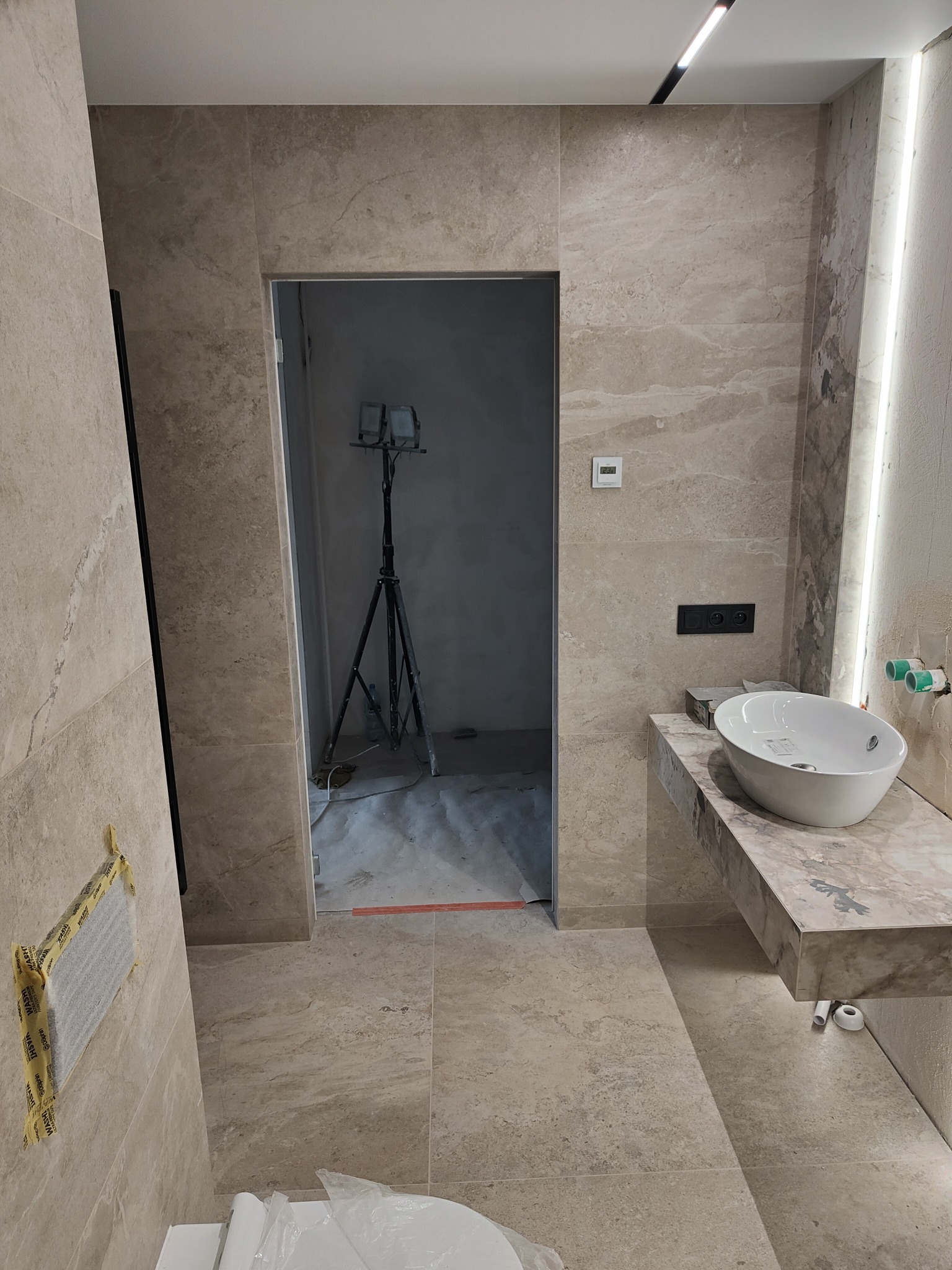 Łazienka w trakcie prac wykończeniowych: beżowe płytki imitujące marmur, umywalka nablatowa, oświetlenie LED, widoczny statyw z lampami i zabezpieczony otwór wentylacyjny.