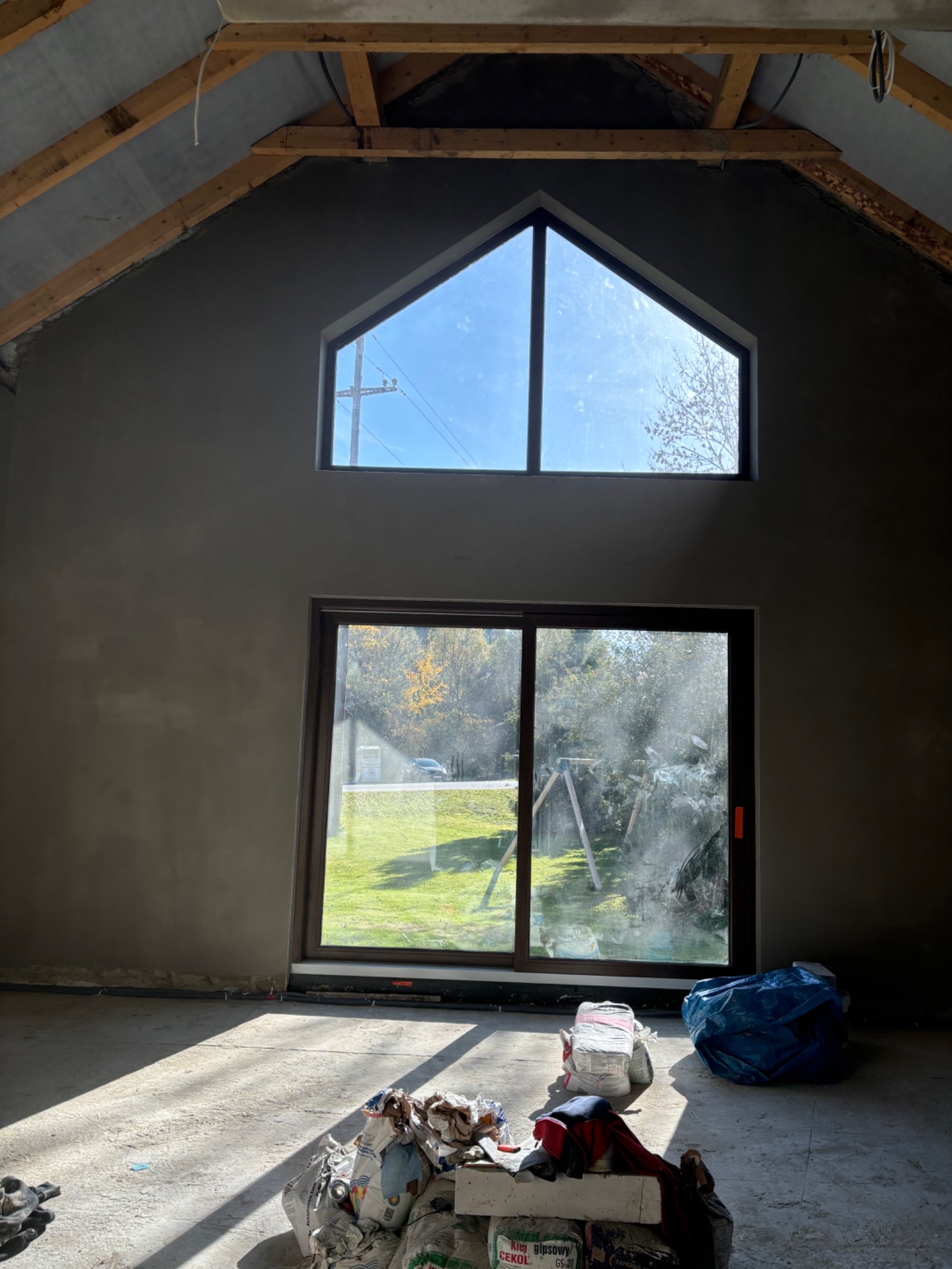 Wnętrze domu w stanie surowym po nałożeniu szarej warstwy wykończeniowej na ściany, widoczne okna z widokiem na zewnątrz, podłoga z resztkami materiałów budowlanych i workami po gipsie.