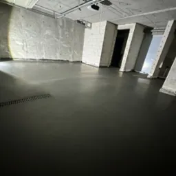 Świeżo wylana, ciemnoszara posadzka z widocznym odpływem liniowym w surowym, betonowym wnętrzu w trakcie prac wykończeniowych.