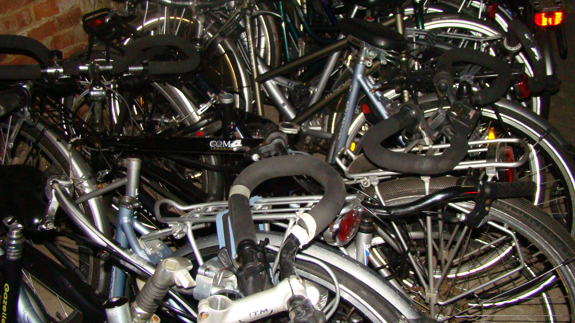 Stłoczone rowery różnych marek, w tym Gazelle, Batavus i Koga Miyata, zaparkowane w pomieszczeniu z ceglaną ścianą w tle.