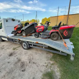 Laweta przewozi dwie kosiarki traktorki marki Faworyt, zabezpieczone pomarańczowymi pasami. Biały samochód na tle zieleni i ogrodzenia z siatki.