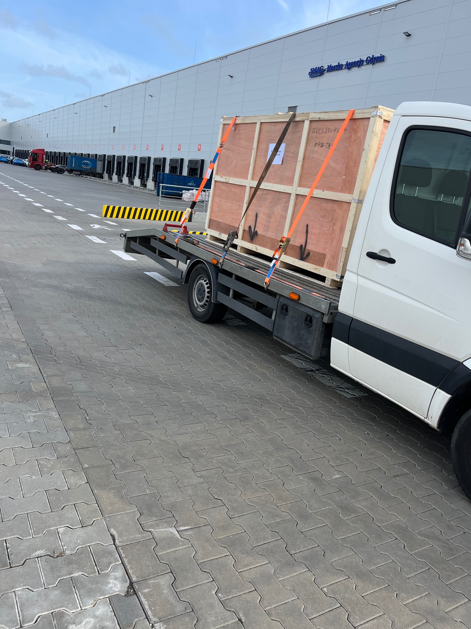 Laweta przewozi zabezpieczoną drewnianą skrzynię na tle magazynu z napisem 'SMAG Morska Agencja Gdynia'.