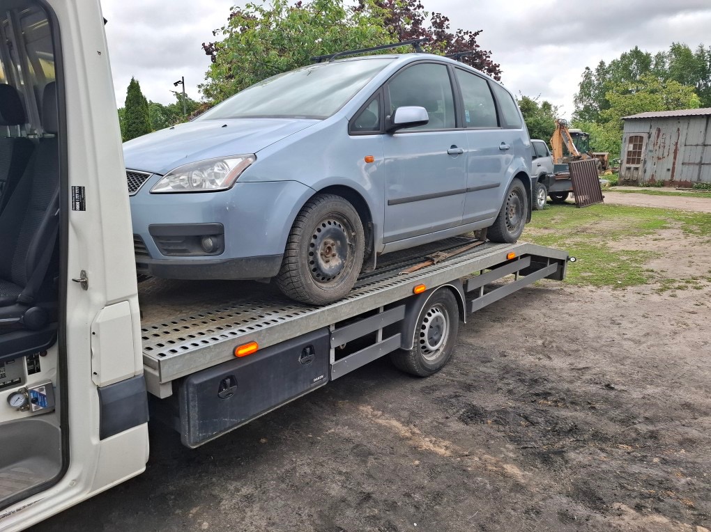 Niebieski Ford C-Max na platformie lawety, widok z boku, lekko z góry. W tle zielone drzewa i budynek gospodarczy. Widoczne opony auta i platformy.