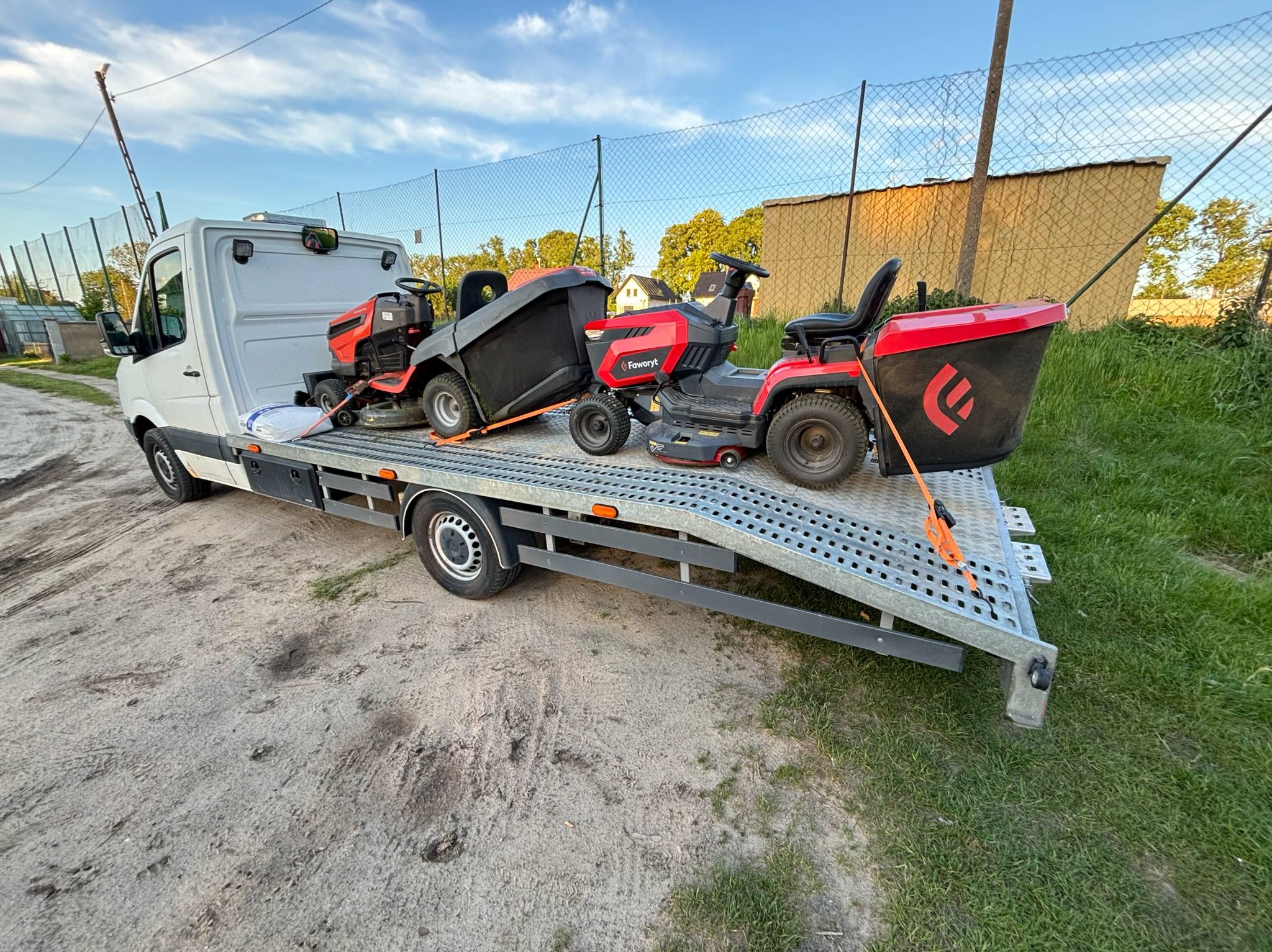 Laweta przewozi dwie kosiarki traktorki marki Faworyt, zabezpieczone pomarańczowymi pasami. Biały samochód na tle zieleni i ogrodzenia z siatki.