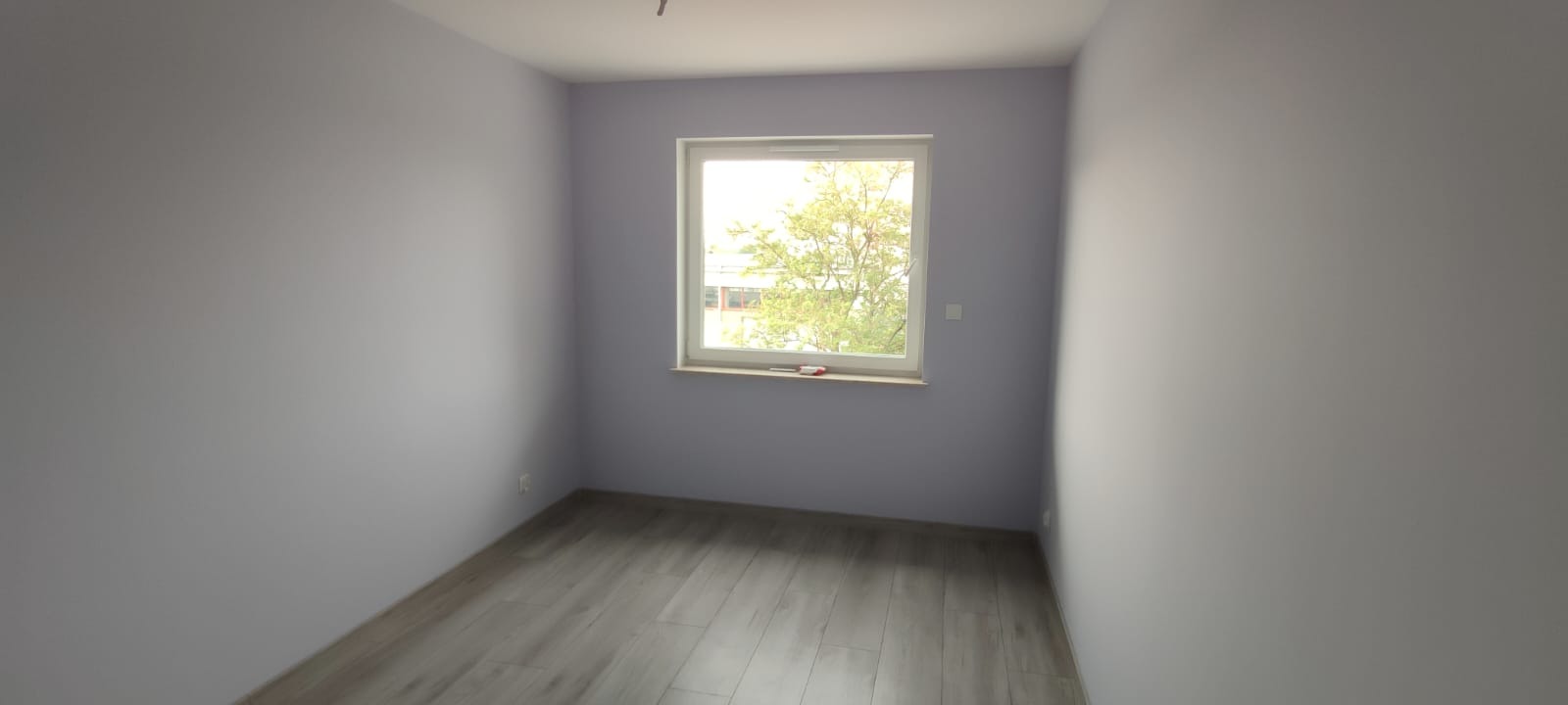 Jasny pokój po odświeżeniu: szare ściany, panele podłogowe, okno z widokiem na drzewo. Na parapecie leży nóż do tapet.