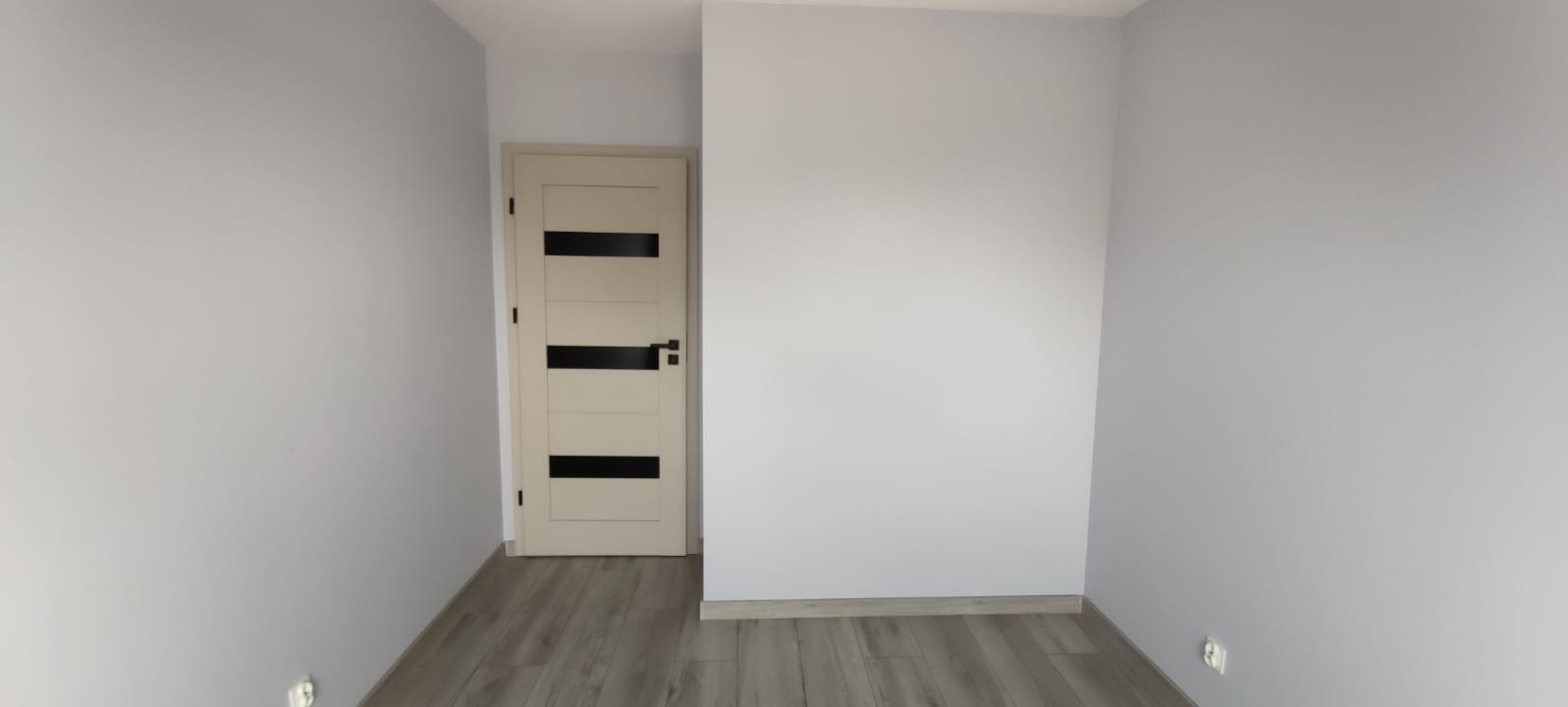 Wnętrze po remoncie: szare ściany, panele podłogowe w odcieniach szarości i beżowe drzwi z czarnymi, poziomymi przeszkleniami. Widok na fragment pokoju.