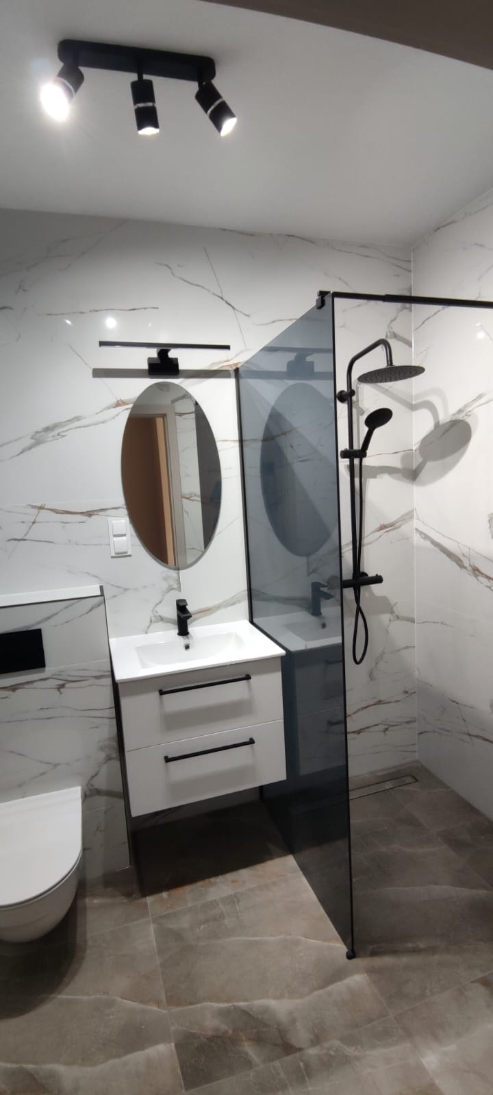 Wykończona łazienka z kabiną typu walk-in, umywalką z szafką i owalnym lustrem. Armatura w kolorze czarnym. Na ścianach i podłodze płytki imitujące marmur.