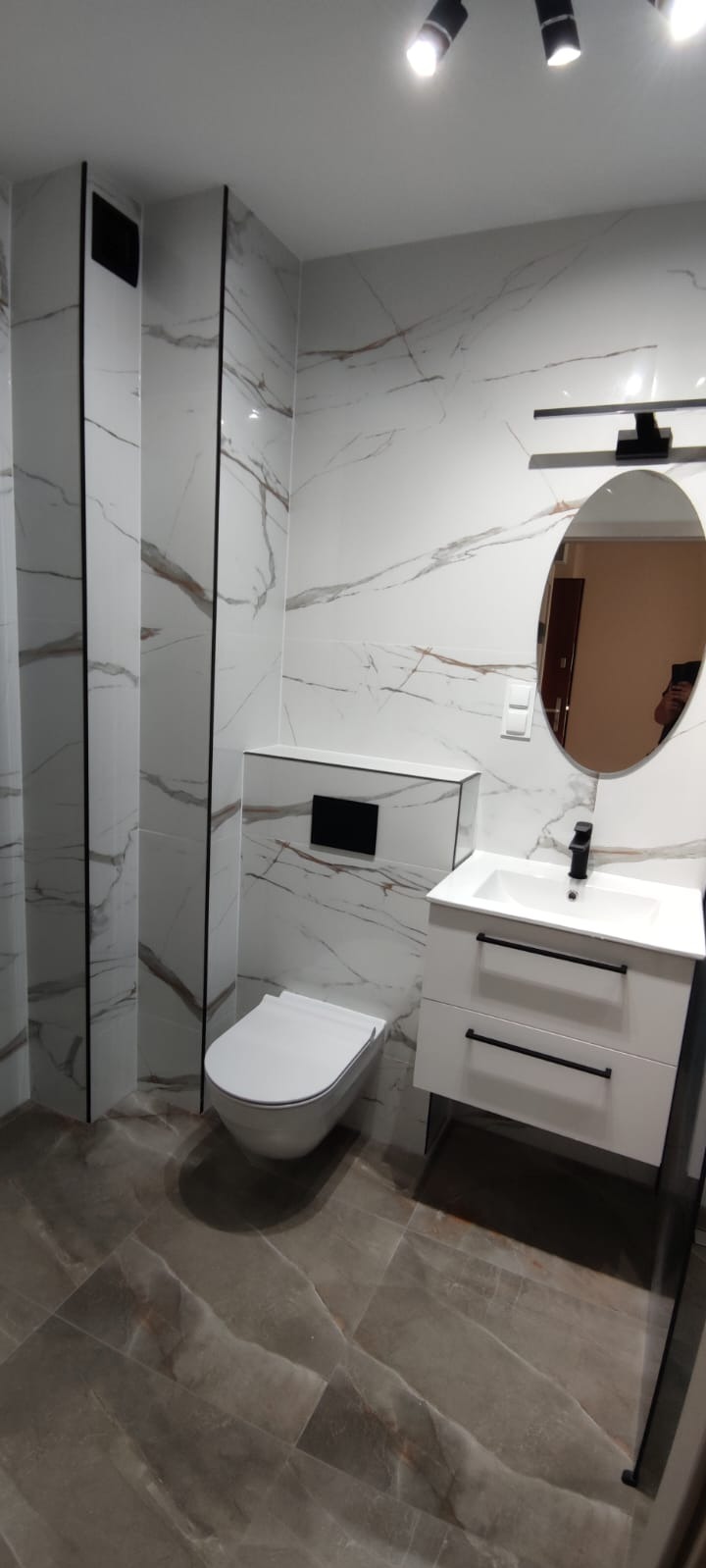 Wykończona łazienka z marmurowymi płytkami, wiszącą toaletą i szafką z umywalką. Czarna armatura i oświetlenie punktowe dodają elegancji.