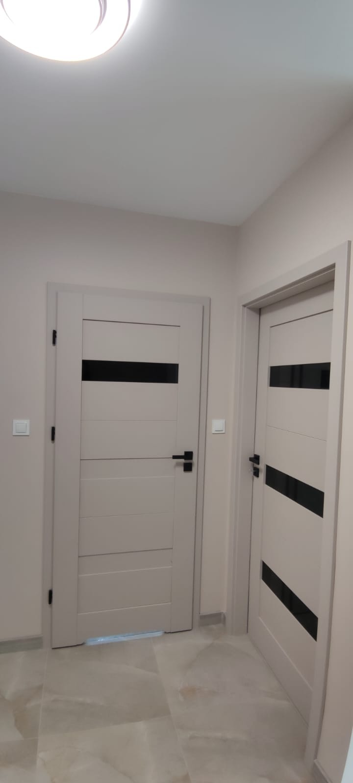 Dwie jasne, nowoczesne drzwi z czarnymi, poziomymi wstawkami w korytarzu z beżowymi ścianami i marmurową podłogą. Czarne klamki i osprzęt.