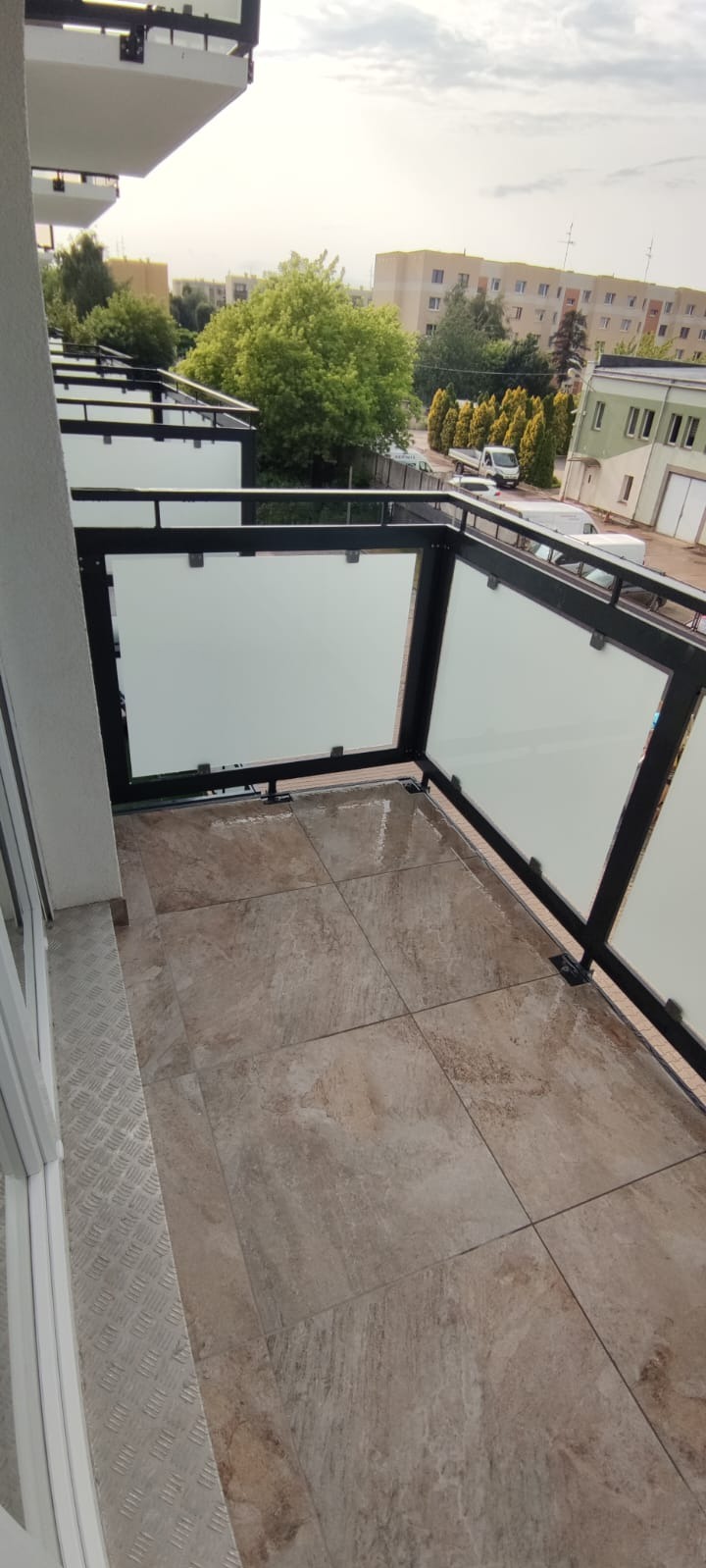 Wykończony balkon z brązowymi płytkami podłogowymi i szklanymi balustradami. Widok na zieleń i budynki w tle. Ujęcie z góry pokazuje detale wykończenia.