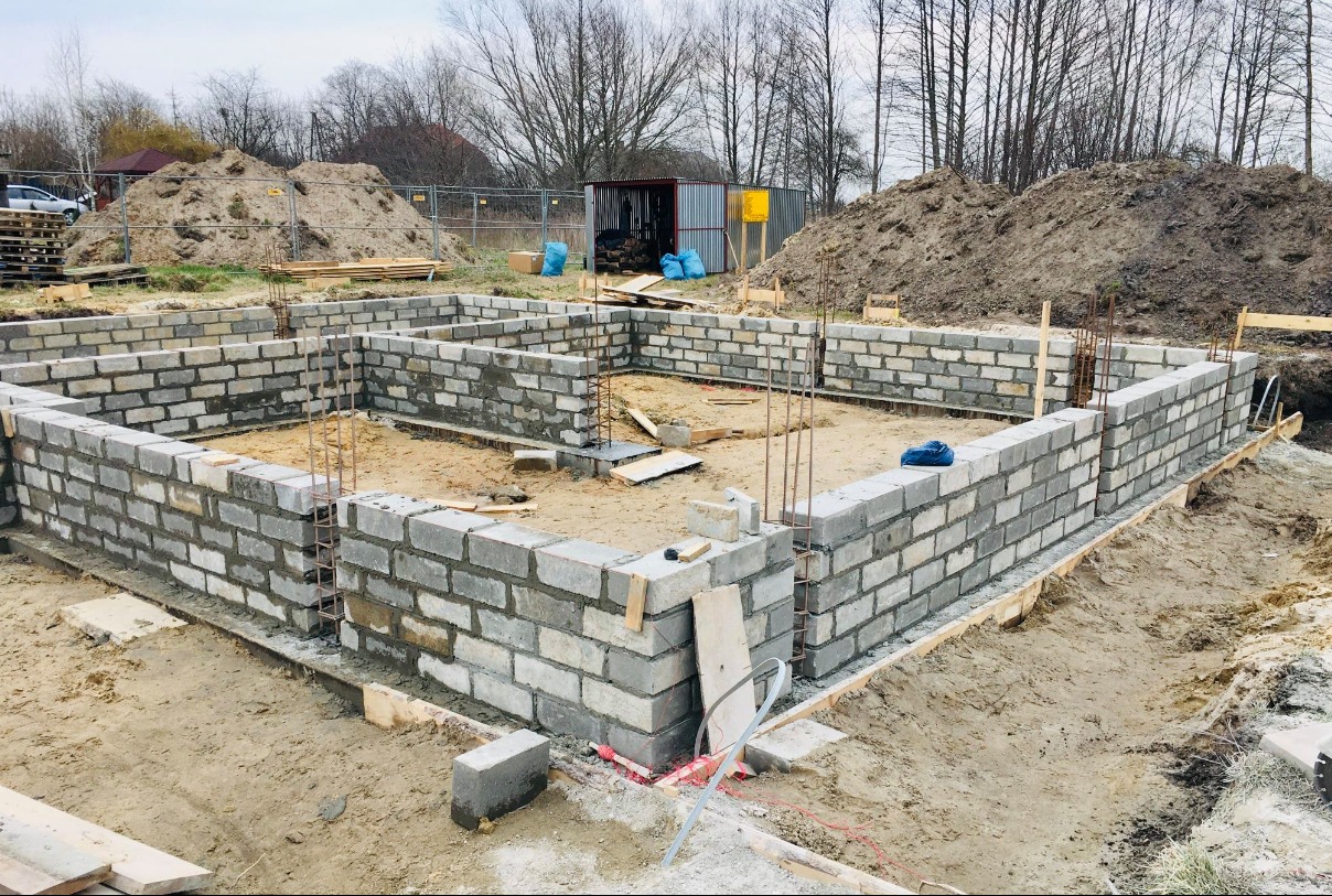 Fundamenty domu jednorodzinnego w trakcie budowy, widoczne bloczki betonowe, wystające pręty zbrojeniowe, w tle sterta ziemi i blaszany barak.
