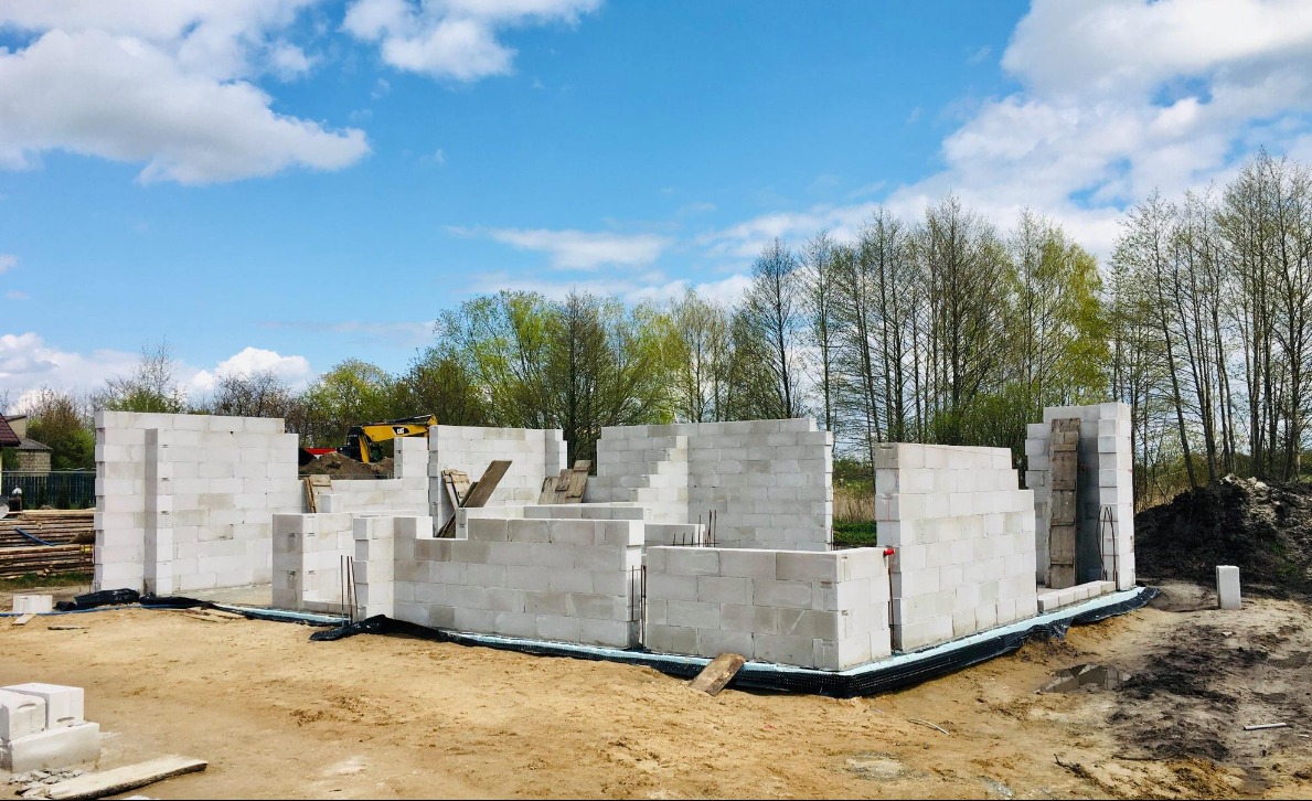 Budowa domu jednorodzinnego w stanie surowym otwartym, ściany z białych bloczków, widoczny żółty sprzęt budowlany w tle, błękitne niebo z chmurami.