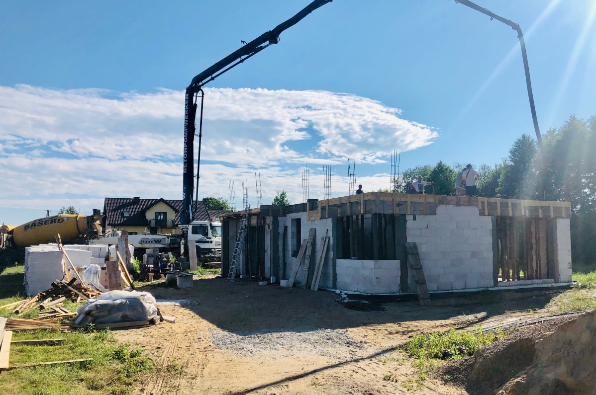 Budowa domu jednorodzinnego w trakcie wznoszenia ścian z betonu komórkowego, z widocznym betonowozem firmy Basero i pompą do betonu na tle błękitnego nieba z chmurami.