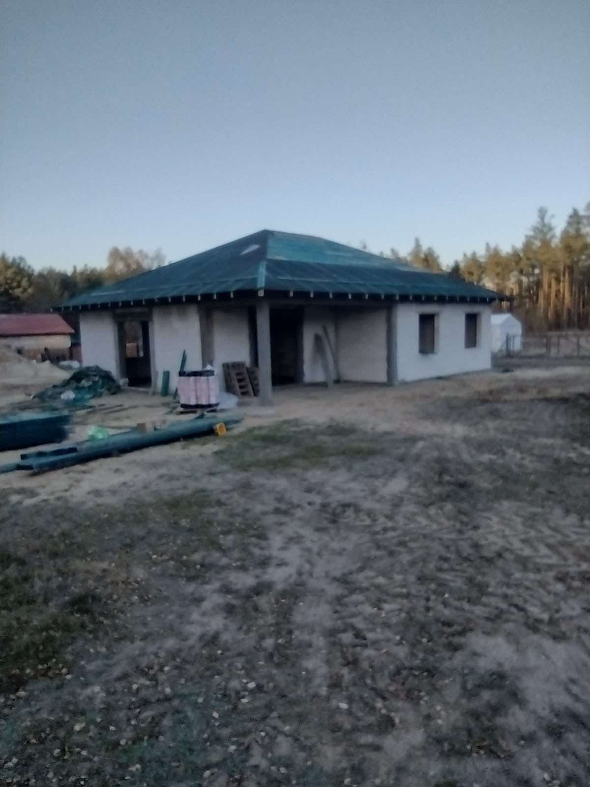 Niedokończony dom jednorodzinny w stanie surowym zamkniętym z ciemnozielonym dachem, widoczne materiały budowlane i otaczający teren.