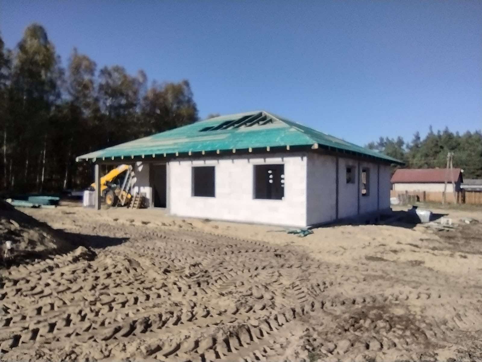 Budowa domu jednorodzinnego w stanie surowym otwartym z widoczną koparkoładowarką i zielonym pokryciem dachu, teren wokół rozjeżdżony przez maszyny budowlane.