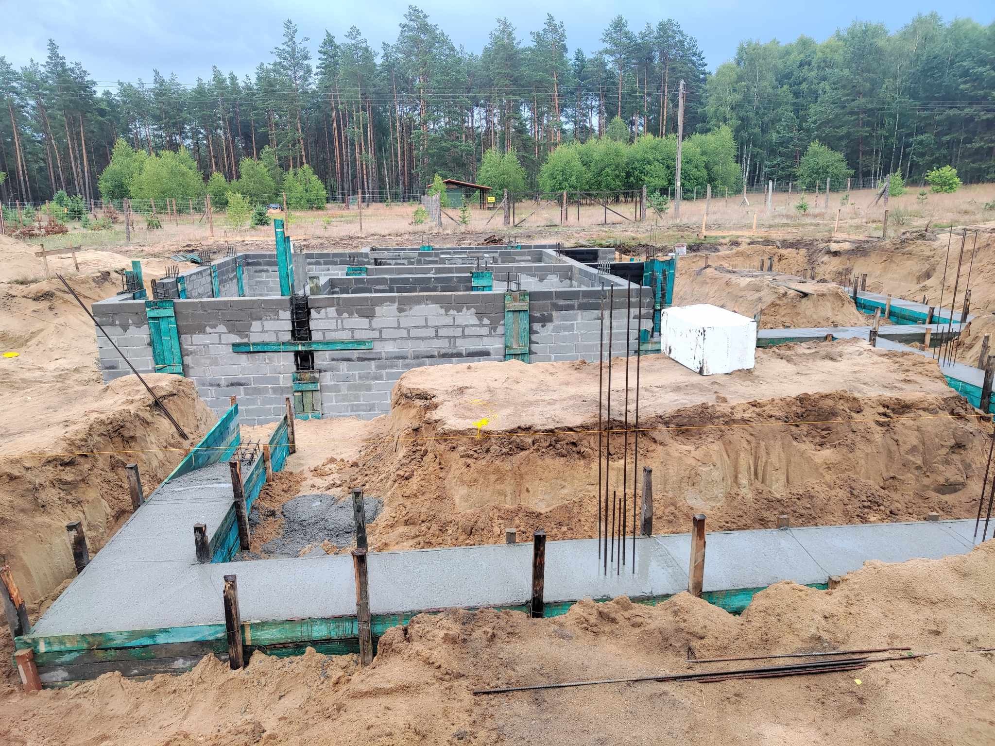 Wykop pod fundamenty domu jednorodzinnego z częściowo wylaną ławą fundamentową i wzniesionymi ścianami fundamentowymi z bloczków betonowych, widoczne szalunki w kolorze turkusowym i wystające pręty...