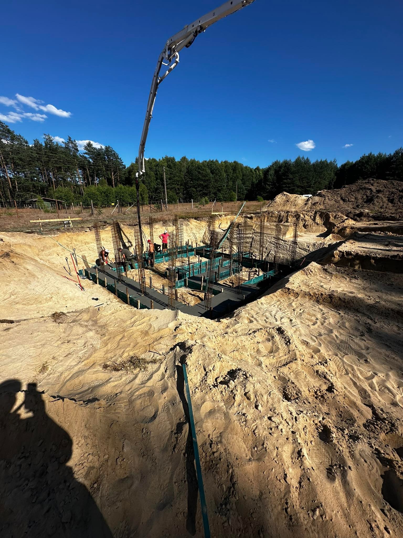 Wykop pod fundamenty domu jednorodzinnego z widocznym zbrojeniem i szalunkiem, w tle las i błękitne niebo, na pierwszym planie piasek i ślady stóp.