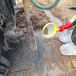 Michał Jarnuczak - Wiertnica w trakcie pracy, osoba w czerwonej rękawicy przesiewa urobek przez żółte sito, widoczny wąż doprowadzający wodę i żwir na ziemi.