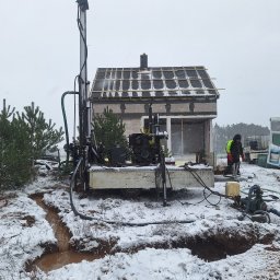 Michał Jarnuczak - Wiercenie studni głębinowej w zimowych warunkach; widoczna wiertnica na przyczepie, fragment niedokończonego budynku w tle i pracownik w odblaskowej kurtce.