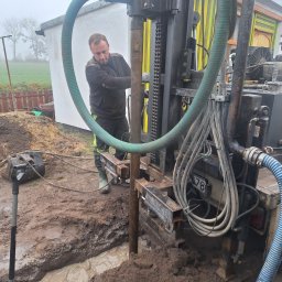 Michał Jarnuczak - Pracownik obsługuje wiertnicę studni głębinowej; widoczna błotnista woda w wykopie, zielone węże i fragment kolorowego budynku w tle.