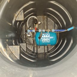 Michał Jarnuczak - Wnętrze czarnego zbiornika z zainstalowanym, niebieskim hydroforem z manometrem, zaworami i przewodami. Widoczne ślady zabrudzeń na ścianach zbiornika.