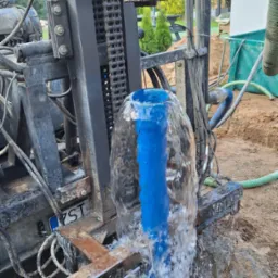 Woda wytryskuje spod maszyny wiertniczej z widoczną tablicą rejestracyjną i fragmentem domu w tle.