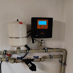 Studniarstwo- studnie głębinowe, odwierty techniczne - Instalacja hydroforowa IBO z czarnym panelem sterującym na białej ścianie. Widoczne rury, zawory i filtr. Element instalacji wodnej w pomieszczeniu.