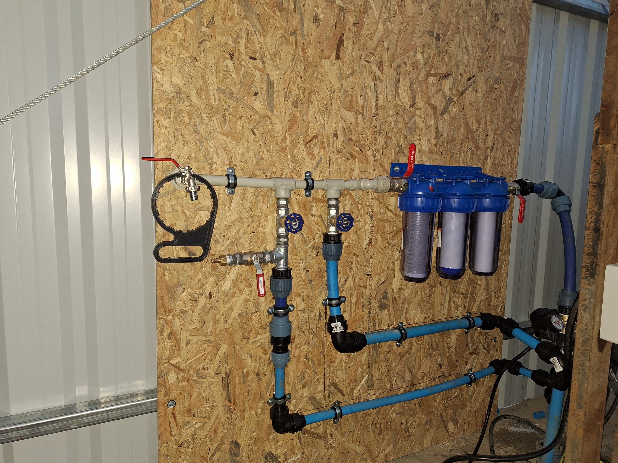 Zmontowany system filtrów wody z niebieskimi obudowami i manometrem, przymocowany do ściany z płyty OSB wewnątrz pomieszczenia.