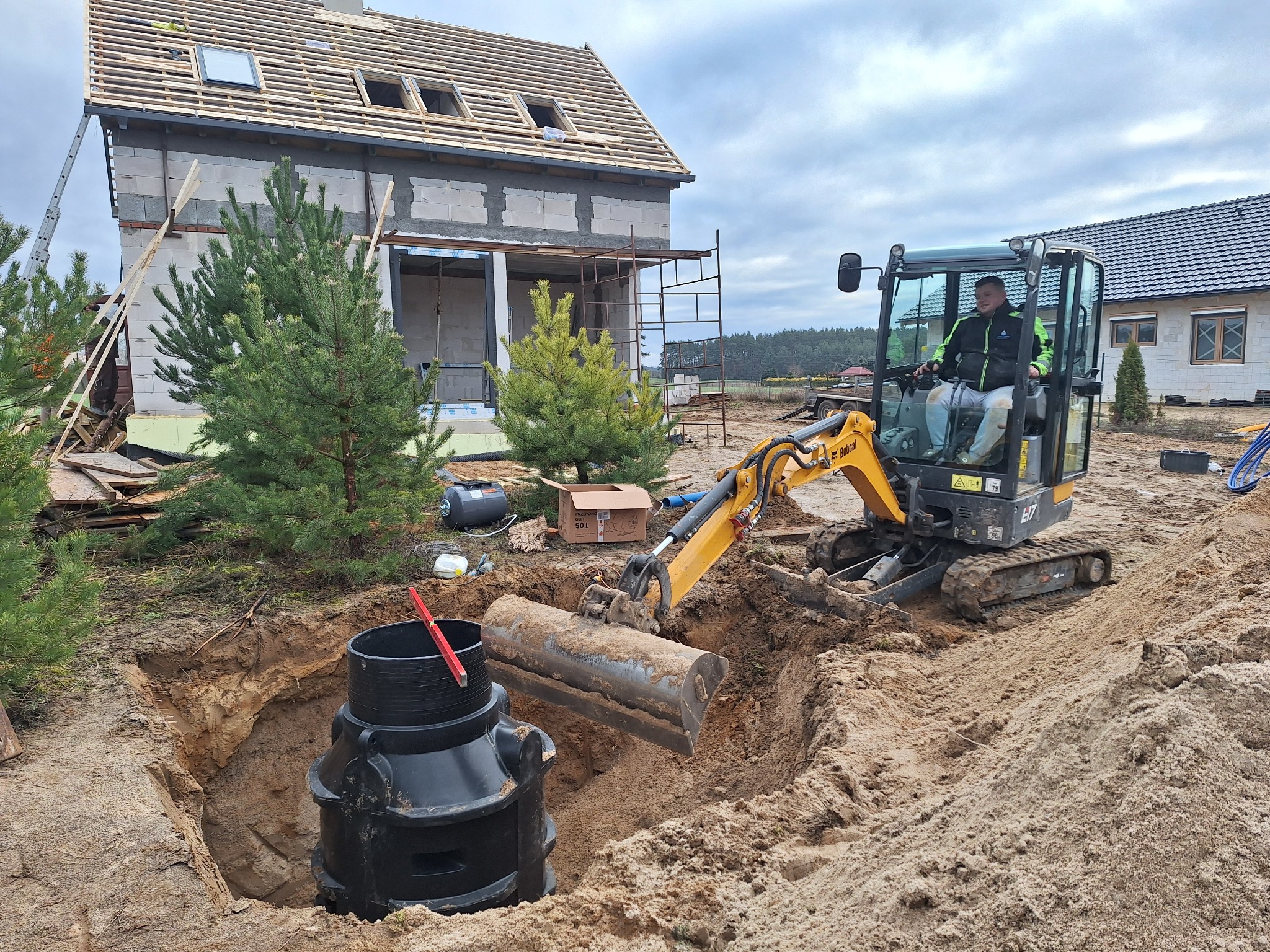 Operator koparki Bobcat w trakcie prac ziemnych przy wykopie pod czarny zbiornik z czerwoną poziomicą, w tle budynek w stanie surowym otwartym z drewnianą konstrukcją dachu i częściowo ocieplony.