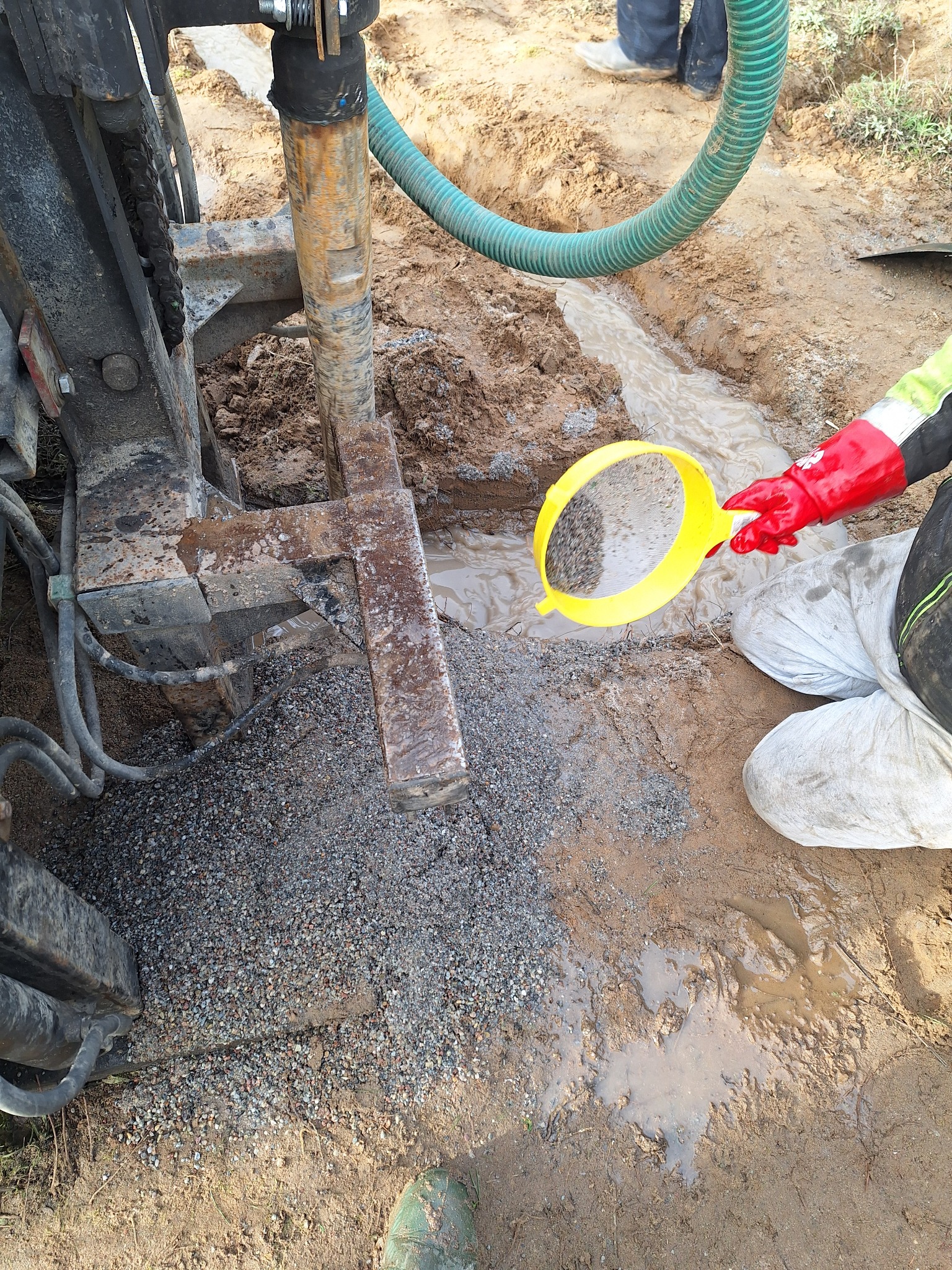 Wiertnica w trakcie pracy, osoba w czerwonej rękawicy przesiewa urobek przez żółte sito, widoczny wąż doprowadzający wodę i żwir na ziemi.