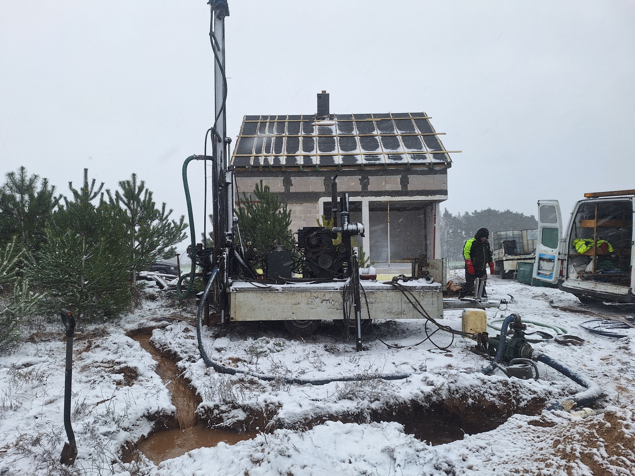 Wiercenie studni głębinowej w zimowych warunkach; widoczna wiertnica na przyczepie, fragment niedokończonego budynku w tle i pracownik w odblaskowej kurtce.