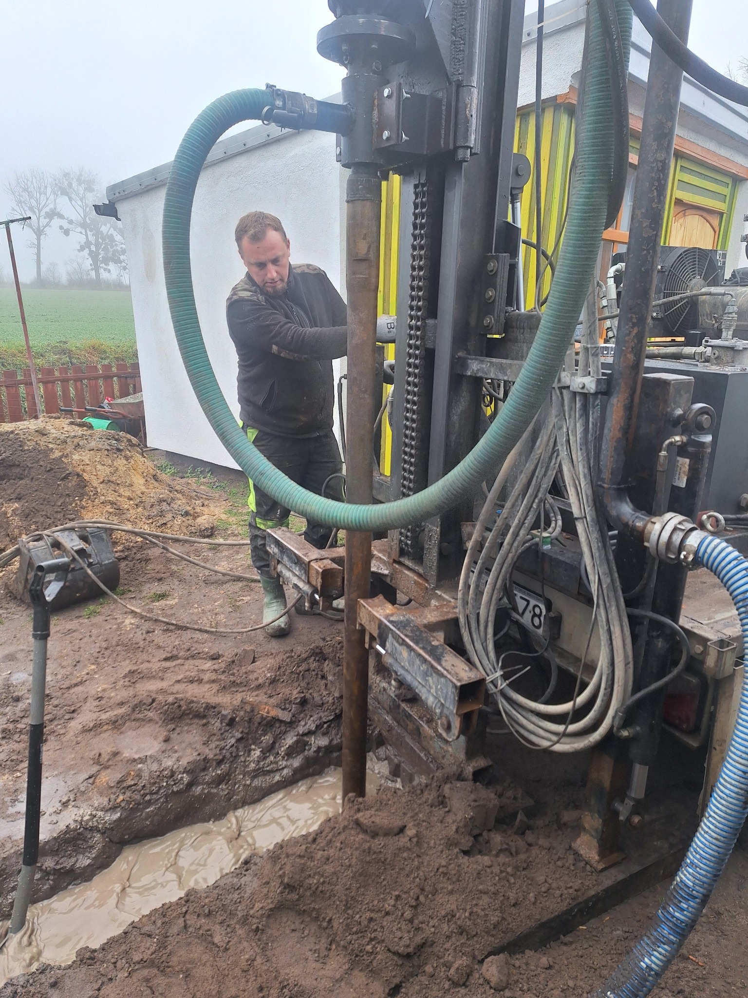 Pracownik obsługuje wiertnicę studni głębinowej; widoczna błotnista woda w wykopie, zielone węże i fragment kolorowego budynku w tle.