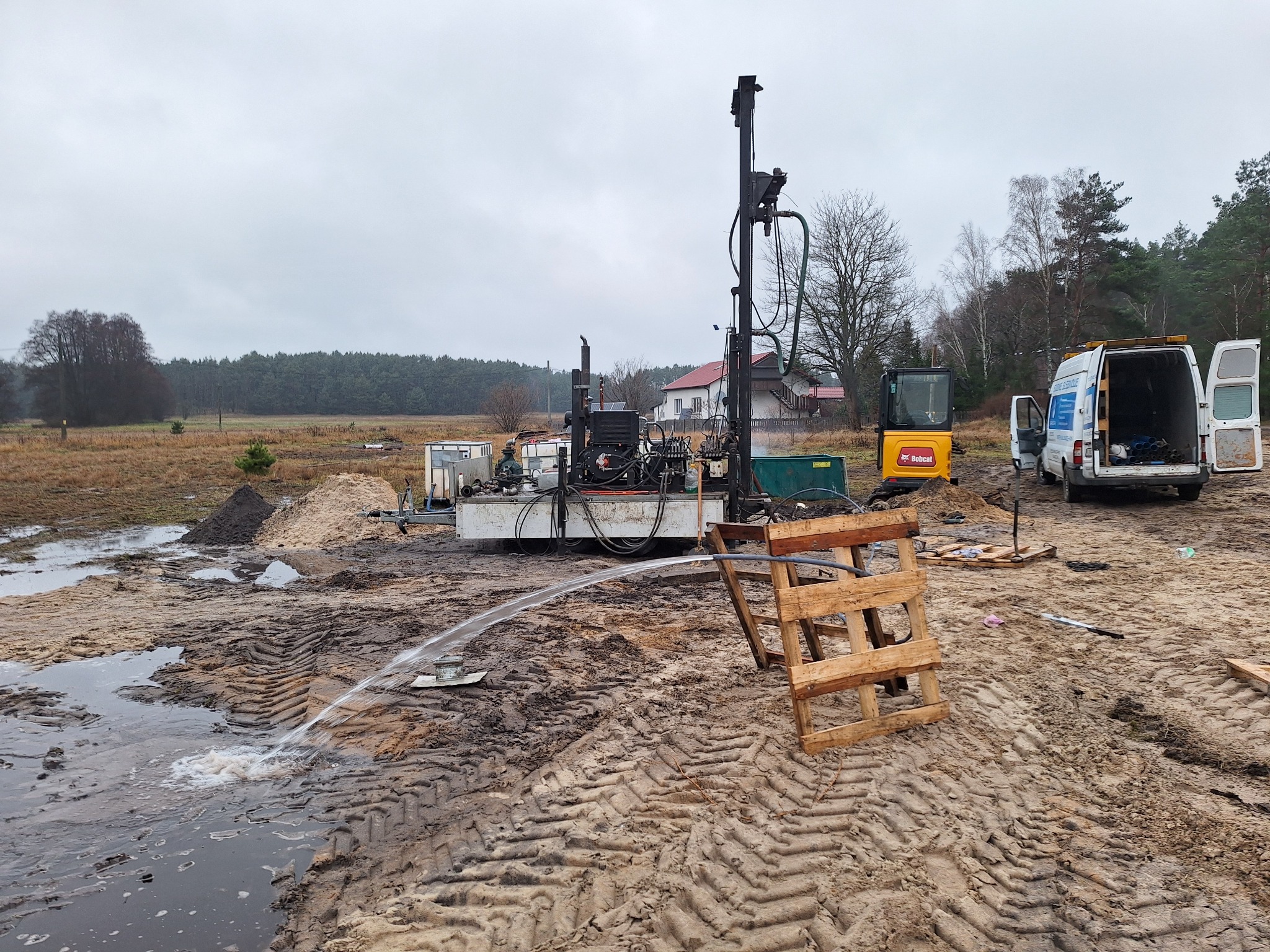Wiertnica do studni głębinowych w trakcie pracy, woda tryska z otworu, widoczny mini-koparka Bobcat i samochód dostawczy na podmokłym terenie.