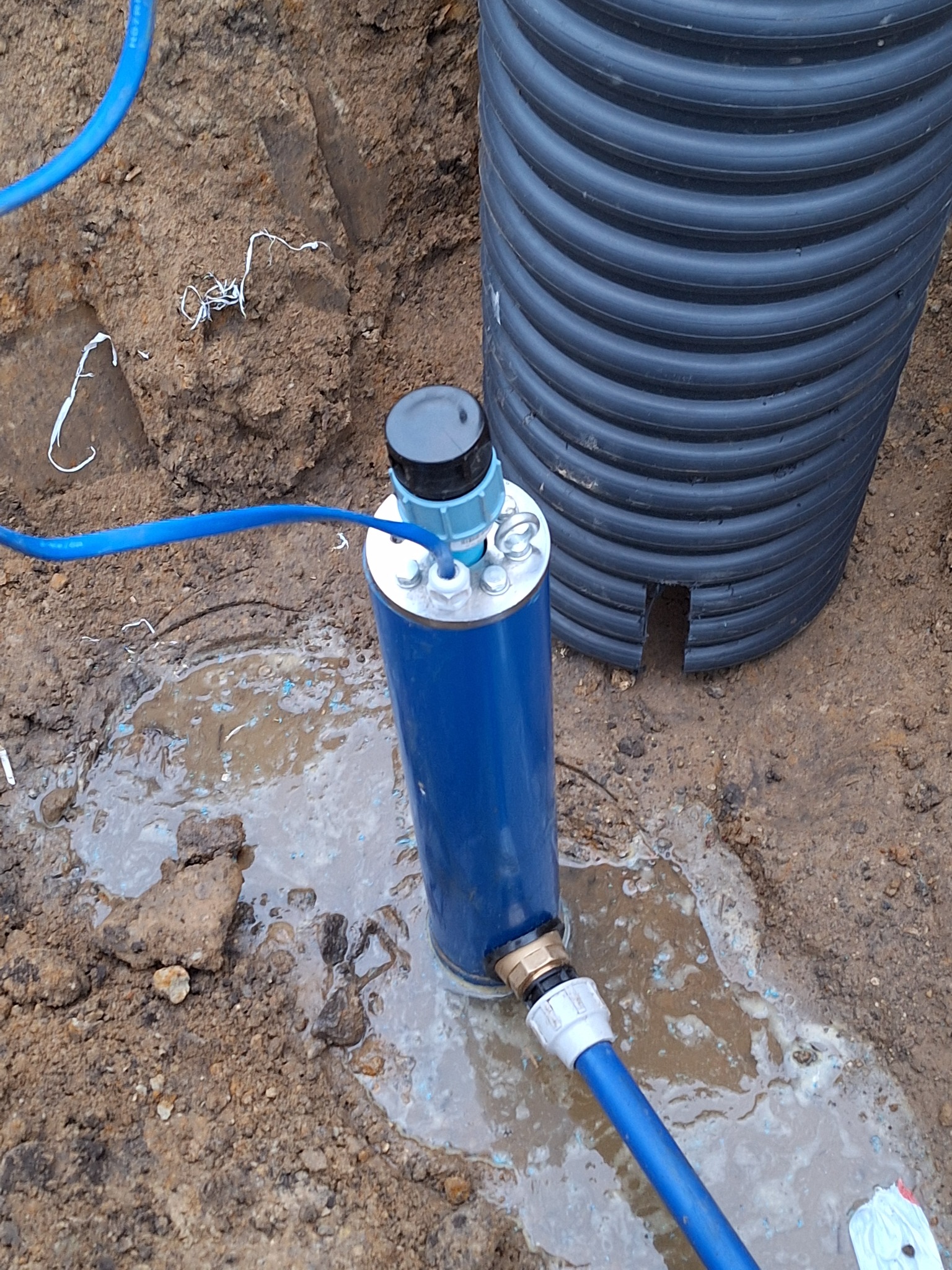 Niebieska rura w wykopie, podłączona do węża, obok karbowanej rury drenażowej; woda w wykopie.
